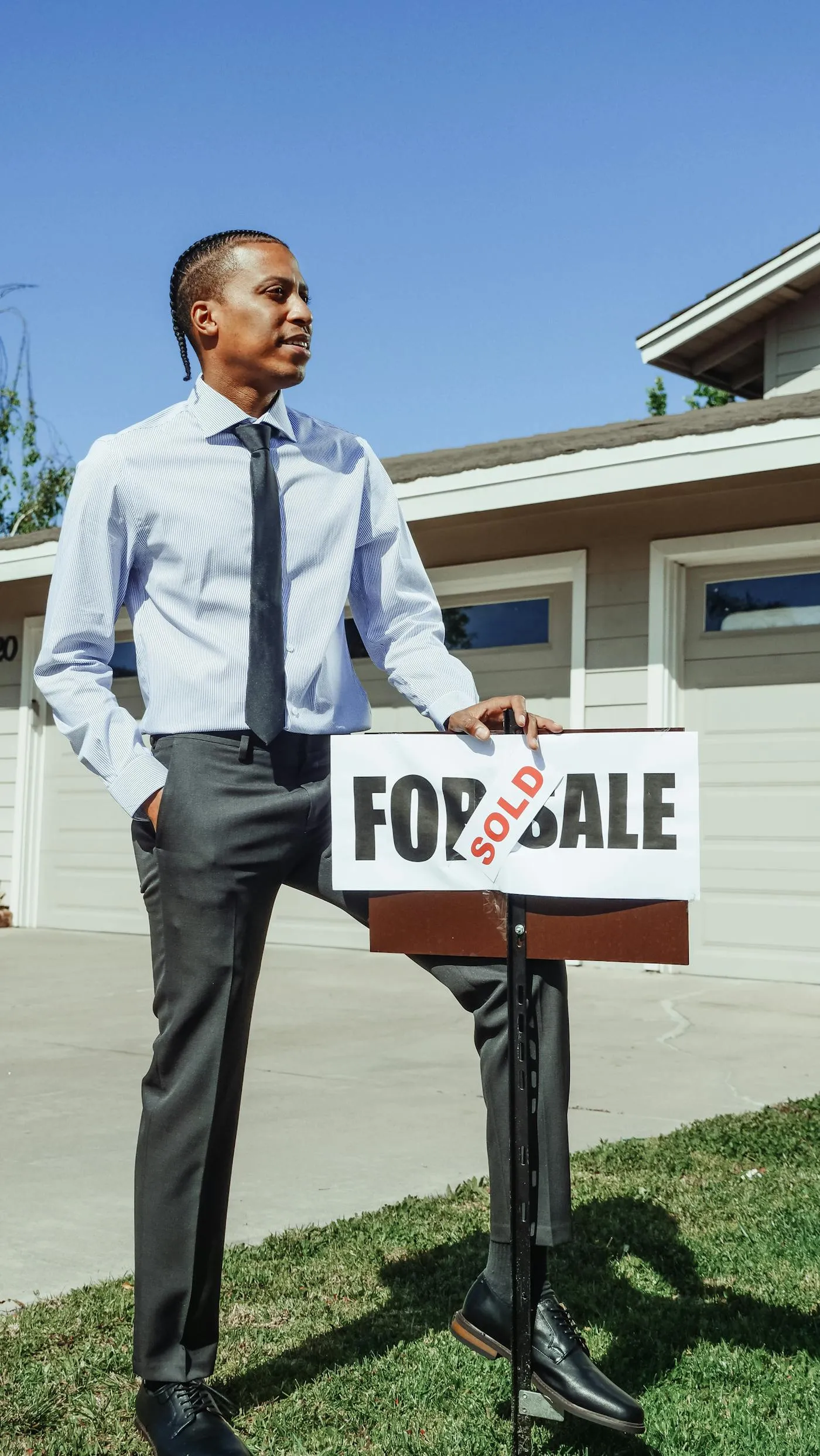 Real-estate agent in blue dress shirt standing beside a sold sign in front of a residential home — pre-sale property context